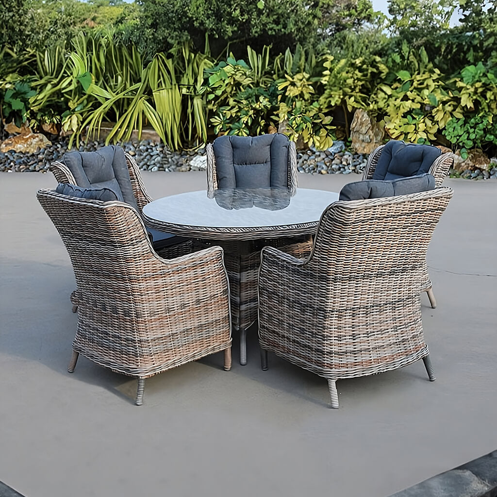 Σετ τραπεζαρίας Naxos από φυσικό ρατάν: στρογγυλό τραπέζι με γυάλινη επιφάνεια, πέντε πολυθρόνες με γκρι καπιτονέ μαξιλάρια και μεταλλικά πόδια, σε αίθριο.