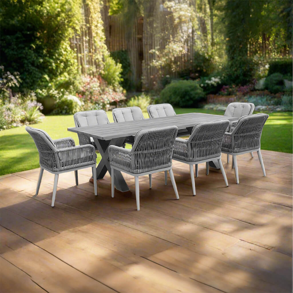 Σετ τραπεζαρίας εξωτερικού χώρου Χαβάη από ανοιχτό γκρι αλουμίνιο: τραπέζι με πόδια Χ και οκτώ γκρι πολυθρόνες με σχοινί και ανοιχτόχρωμα μαξιλάρια, σε ξύλινο κατάστρωμα κήπου. | Lux Furniture