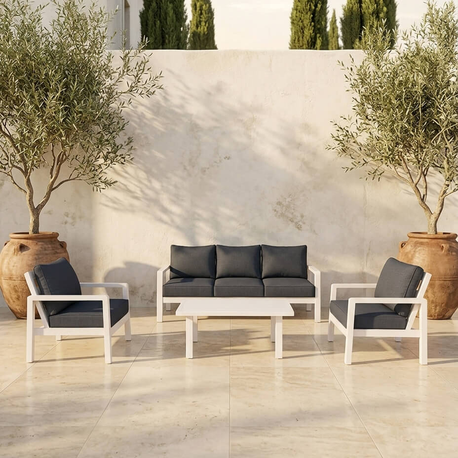 Σετ σαλονιού εξωτερικού χώρου με λευκό ματ σκελετό αλουμινίου και σκούρα γκρι μαξιλάρια, περιλαμβάνει τριθέσιο καναπέ, δύο πολυθρόνες και τραπεζάκι, τοποθετημένο σε αυλή μοντέρνου σπιτιού με γρασίδι και ψηλά δέντρα στο φόντο. / White