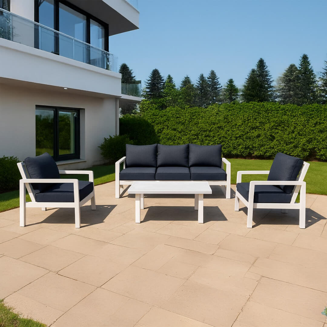 Σετ σαλονιού εξωτερικού χώρου με λευκό ματ σκελετό αλουμινίου και σκούρα γκρι μαξιλάρια, περιλαμβάνει τριθέσιο καναπέ, δύο πολυθρόνες και τραπεζάκι, τοποθετημένο σε αυλή μοντέρνου σπιτιού με γρασίδι και ψηλά δέντρα στο φόντο. / White