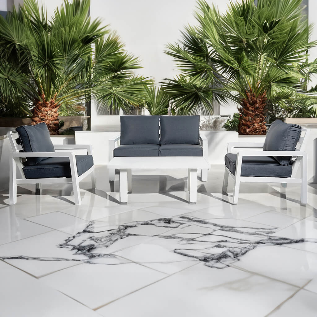Σετ εξωτερικού χώρου Kriti με λευκό ηλεκτροστατικά βαμμένο αλουμινένιο σκελετό και σκούρα γκρι μαλακά μαξιλάρια καθίσματος και πλάτης, που περιλαμβάνει δύο πολυθρόνες, διθέσιο καναπέ και ορθογώνιο τραπεζάκι, τοποθετημένο σε λευκή μαρμάρινη βεράντα με γλάστρες φοίνικα στο φόντο. | Lux Furniture / White