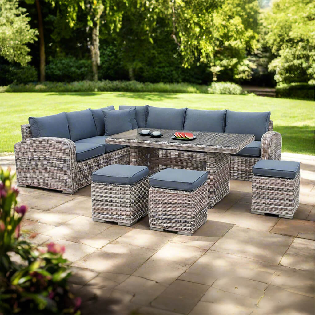 Σετ εξωτερικού χώρου Tilos από ραττάν σε ανοιχτό γκρι με γκρι μαξιλάρια καθίσματος και πλάτης, που περιλαμβάνει γωνιακό καναπέ σε σχήμα Γ, τρία ταιριαστά σκαμπό και ορθογώνιο τραπέζι με γυάλινη επιφάνεια, τοποθετημένο σε πλακόστρωτη αυλή με χλοοτάπητα και δέντρα στο φόντο. | Lux Furniture