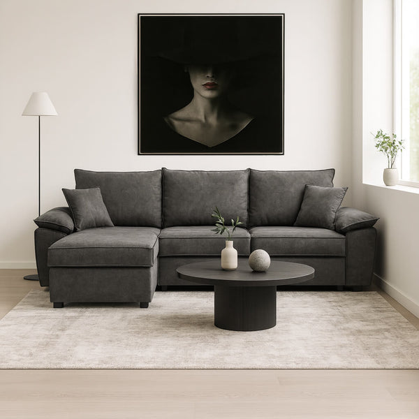 A large, dark grey corner sofa with a left-hand corner (chaise longue) in a minimal living room. The sofa has plush cushions and pairs with a round, dark coffee table. On the white wall behind it hangs a striking, large artwork depicting a woman in a hat that casts a shadow over her face. / Grey