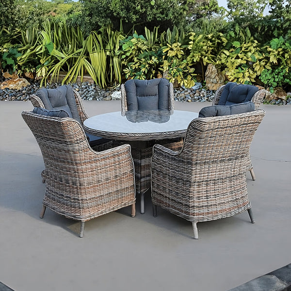 Dining set Naxos from natural rattan: round table with glass top, five armchairs with grey tufted cushions and metal legs, in an atrium.