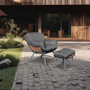 Syros outdoor armchair with a metal frame finished in light grey and orange rattan, with a dark grey buttoned cushion and matching footstool on stone paving, in front of a modern wooden house, surrounded by greenery.