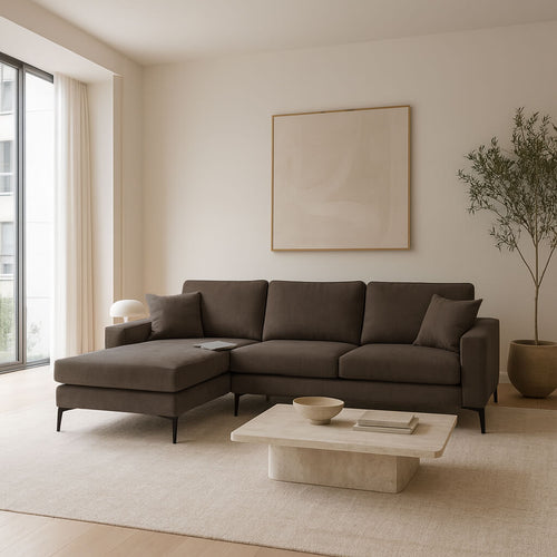 A modern brown corner sofa with a left corner (chaise longue) and slim black metal legs that create a light, raised feel. It sits in a bright, airy living room with a neutral colour palette, and is paired with a square travertine coffee table, a large potted olive tree and minimal wall art. / Brown