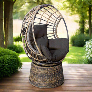 Woven rattan garden swing armchair in brown and grey tones with dark grey seat and back cushions, on a round rattan base on a wooden veranda, with trees and shrubs in the background. | Lux Furniture