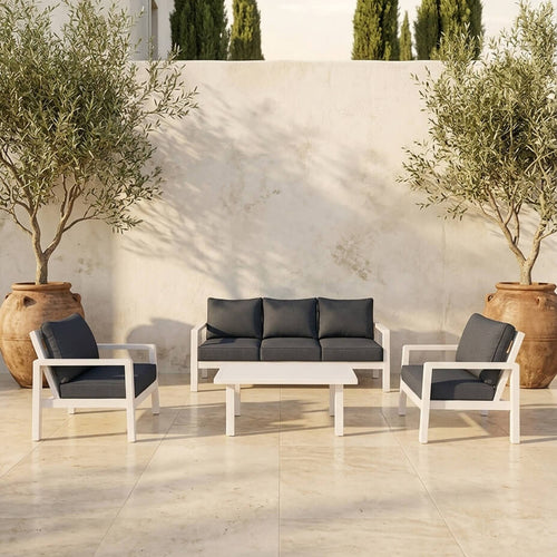Outdoor lounge set with a matte white aluminium frame and dark grey cushions, including a three-seater sofa, two armchairs and a coffee table, set in the yard of a modern house with grass and tall trees in the background. / White