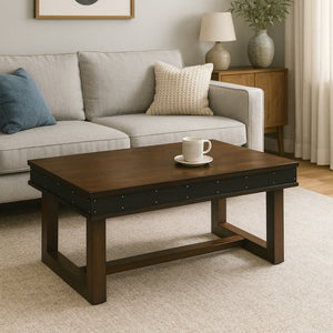 The industrial-style coffee table with a dark wooden top and a metal band with rivets, photographed in a decorated living room. It sits on a beige rug, in front of a comfortable light grey sofa, showing its dimensions and style within a home setting.